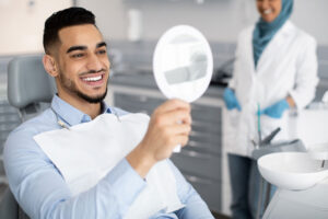 smiling man sits in a dental exam chair holding a mirror as a dental professional explains the benefits of veneers