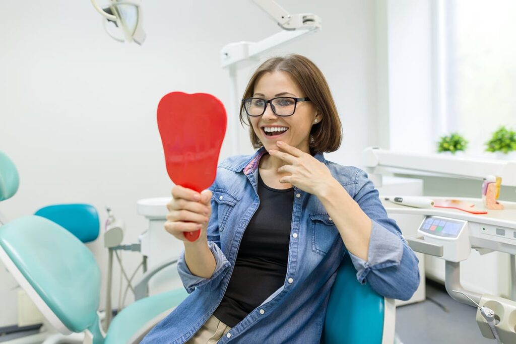 a woman sits in dental exam chair with a mirror towards her admiring her smile and replying yes to the question of are dental implants worth the money