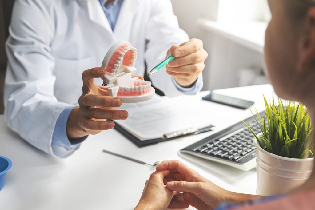 a dentist sits at his desk with a patient showing her dental implants procedure on a mouth model and answering her question about how long do dental implants last