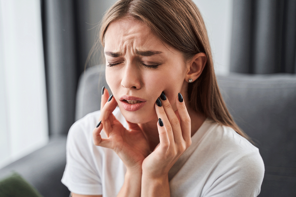 a young woman sits with both hands holding the sides of her face and in pain wondering what to do in a dental emergency