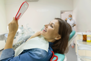 a woman patient experiencing the positive effects of dental implants smiles while looking at herself in a mirror at the dentist office